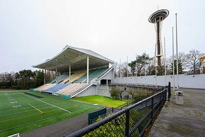 mutilevel grandstands with an angled roof - part of an athletic field below it and a wall behind it. The Space Needle is in  the background behind the wall