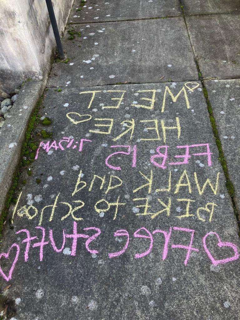 Sidewalk chalk that says "Meet here; Feb 15, Walk and bike to school; Free stuff"