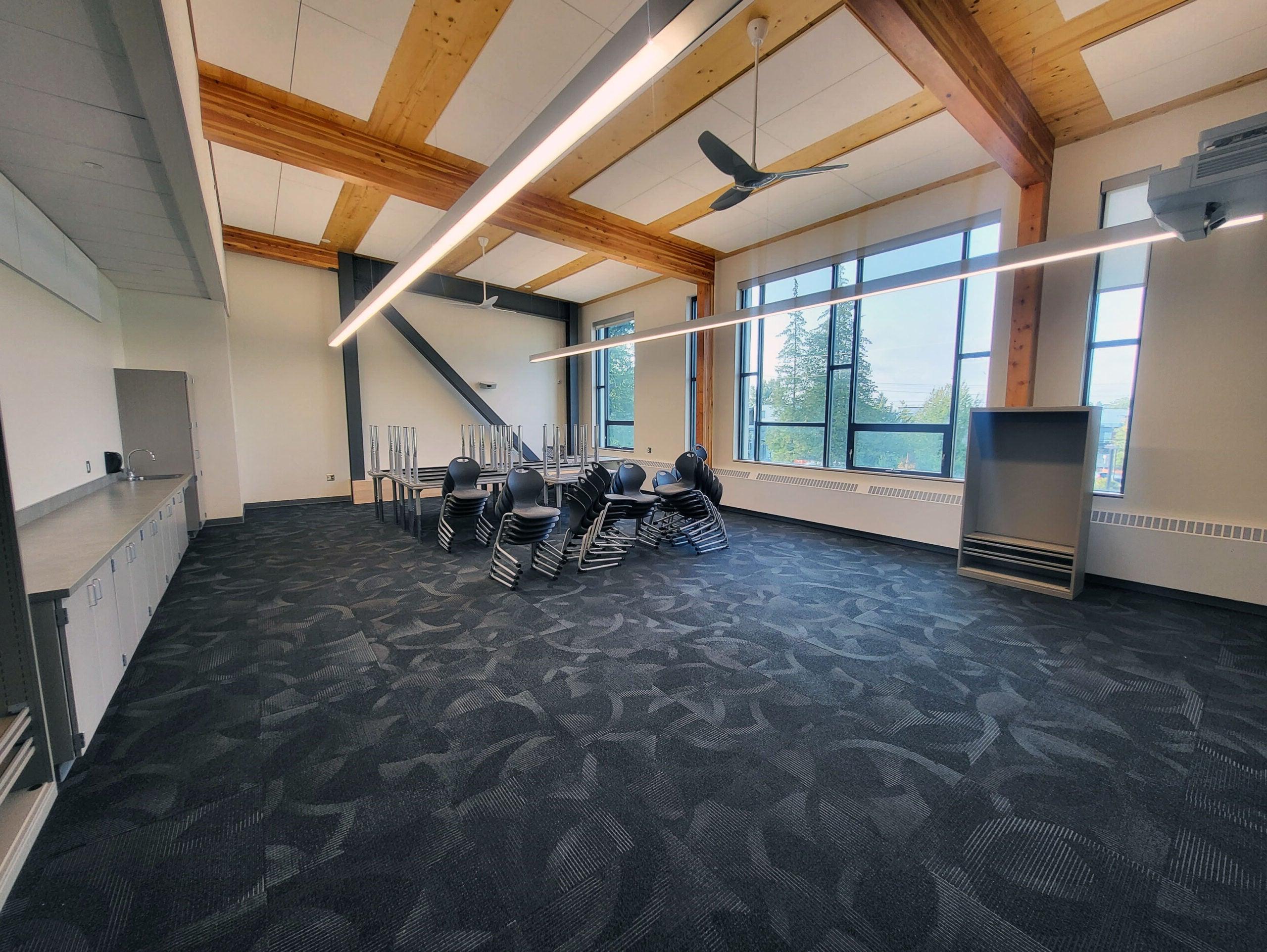 a classroom has windows on one side, wood beams in the ceiling, and a counter across from the windows - chairs and tables are stacked in the center